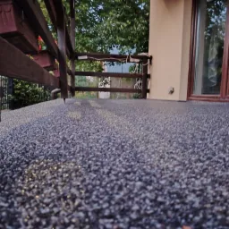 Szara, drobnoziarnista nawierzchnia balkonu z drewnianą balustradą, widok z niskiej perspektywy, z elementami roślinności w tle.