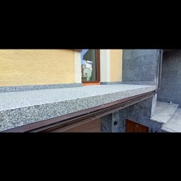 REMO-POS Damian Ostrowski - Wykończony balkon z szarą, kamienną posadzką, brązową ramą i widokiem na okno odbijające budynek. Elewacja budynku w odcieniach beżu i szarości.