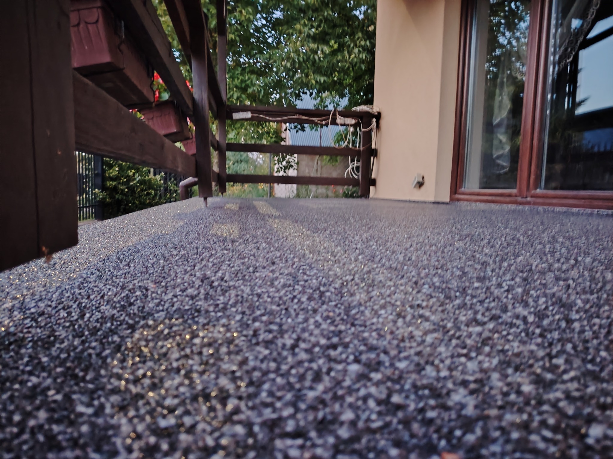 Szara, drobnoziarnista nawierzchnia balkonu z drewnianą balustradą, widok z niskiej perspektywy, z elementami roślinności w tle.