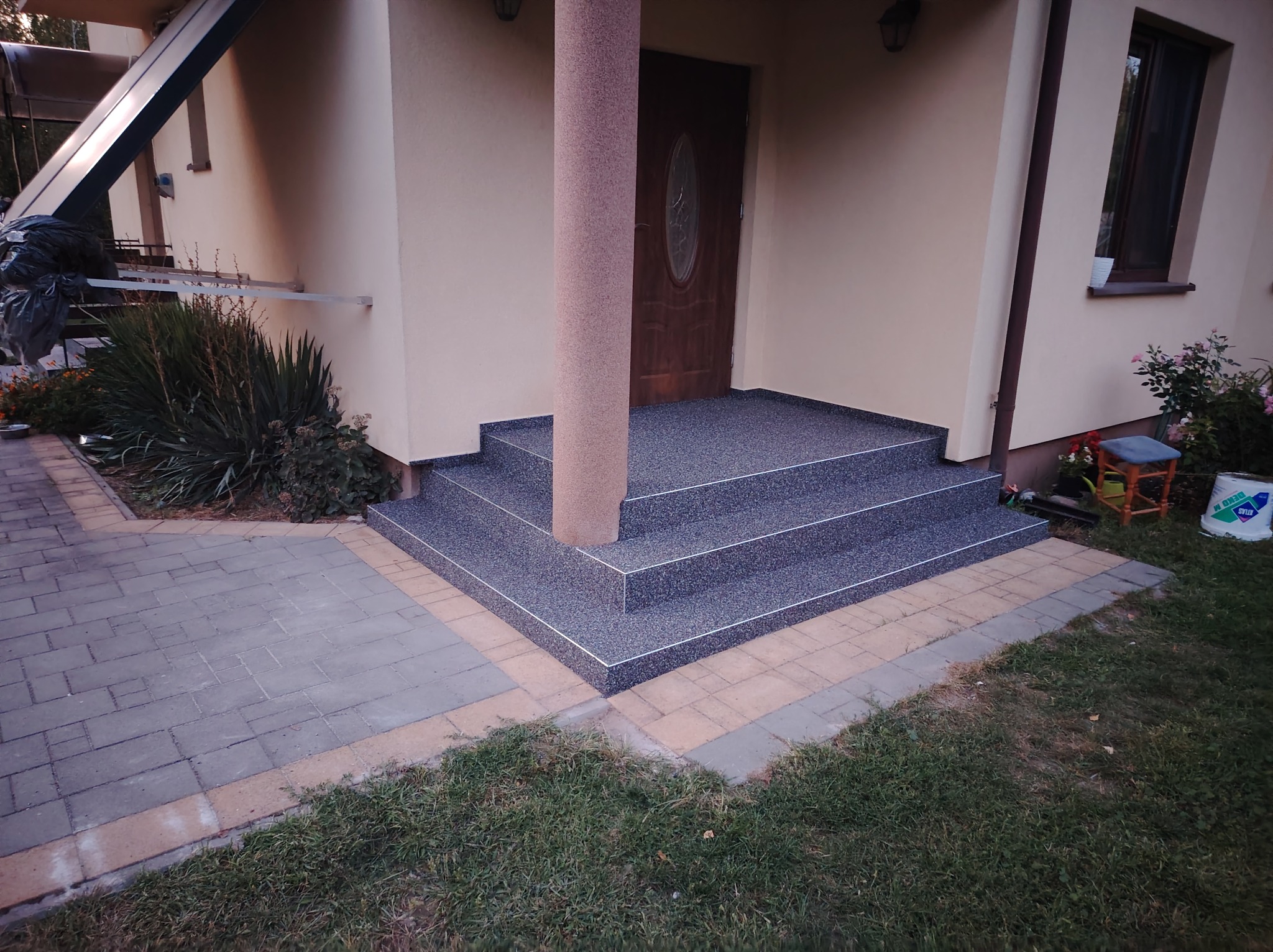 Wejście do domu z trzema schodami pokrytymi szarą, drobnoziarnistą posadzką, z kolumną w tym samym wykończeniu, otoczone kostką brukową i trawnikiem.