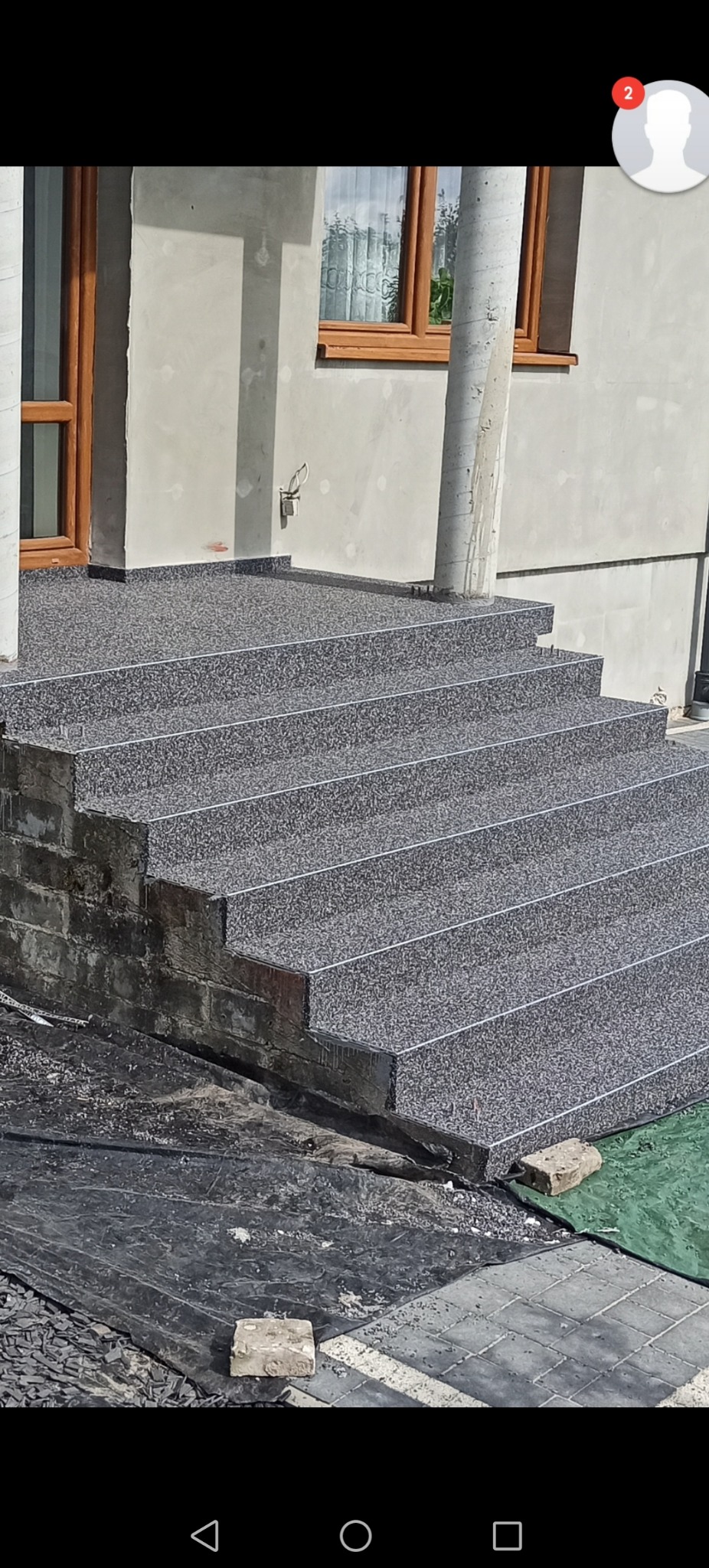 Zewnętrzne schody wejściowe z szarą, drobnoziarnistą nawierzchnią, wykończone listwami, prowadzące do domu z oknem w drewnianej ramie, widoczne ślady prac budowlanych.