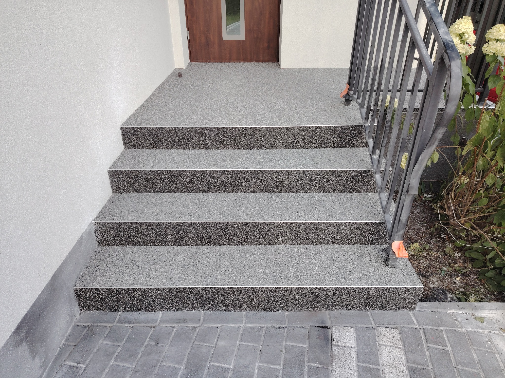 Zewnętrzne schody wejściowe z czterema stopniami wykończone szarą posadzką z żywicy epoksydowej, z metalową barierką po prawej stronie i kostką brukową u podstawy.