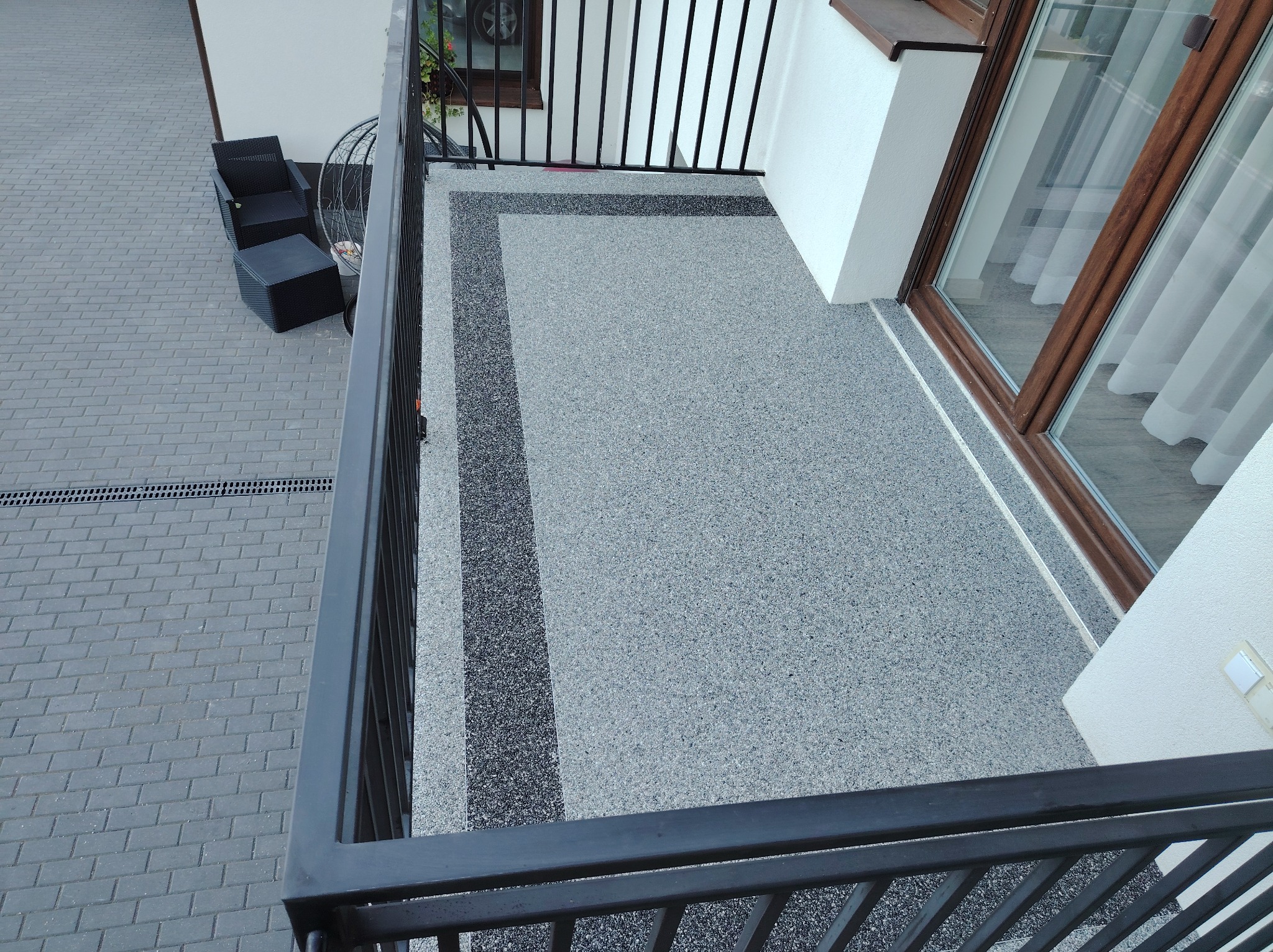 Szary balkon z żywicy epoksydowej z ciemnymi pasami dekoracyjnymi, widok z góry na reling, z meblami ogrodowymi w tle.