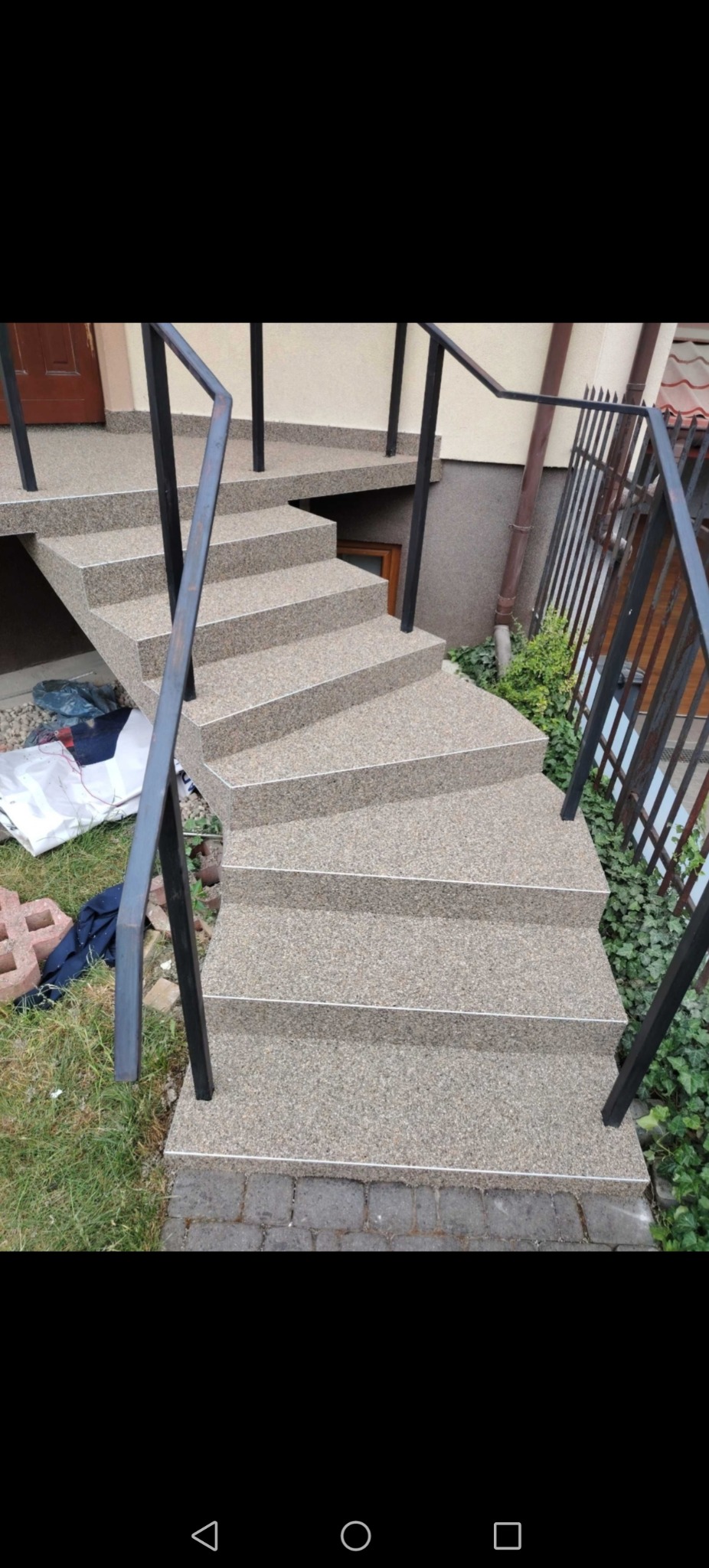 Zewnętrzne schody z szarego kamienia z czarną metalową balustradą prowadzące do drzwi wejściowych, widoczne elementy wykończeniowe i otoczenie z zielenią.