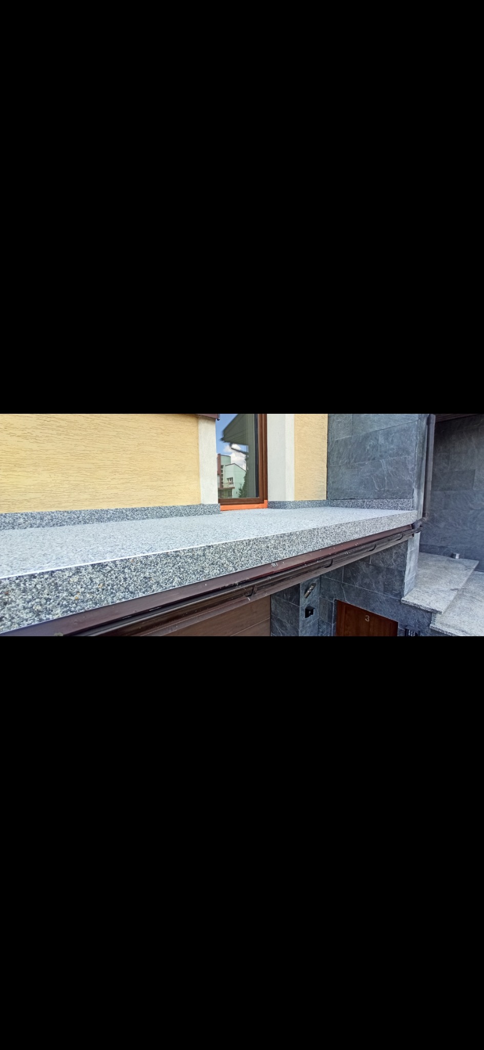 Wykończony balkon z szarą, kamienną posadzką, brązową ramą i widokiem na okno odbijające budynek. Elewacja budynku w odcieniach beżu i szarości.