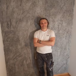 Feduniewicz Bud&Art Wojciech Feduniewicz - Mężczyzna w roboczych spodniach z plamami farby stoi na tle ściany z szarą, strukturalną dekoracją przypominającą kamień.