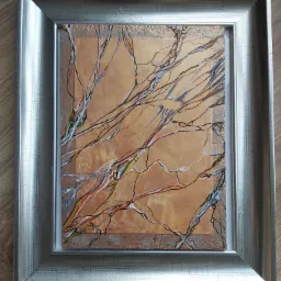 Abstrakcyjny obraz z motywem gałęzi na pomarańczowym tle, oprawiony w srebrną ramę, leżący na drewnianej podłodze.