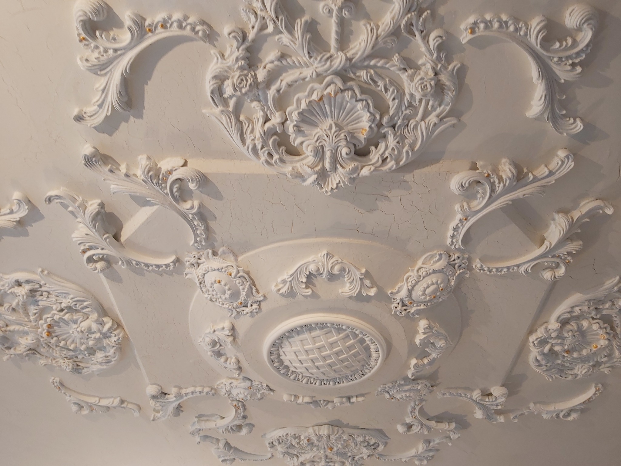 Biały sufit z dekoracyjnymi, gipsowymi ornamentami w stylu klasycznym, z widocznymi drobnymi pęknięciami na powierzchni. Centralny element dekoracyjny przypomina rozetę z motywem plecionki.