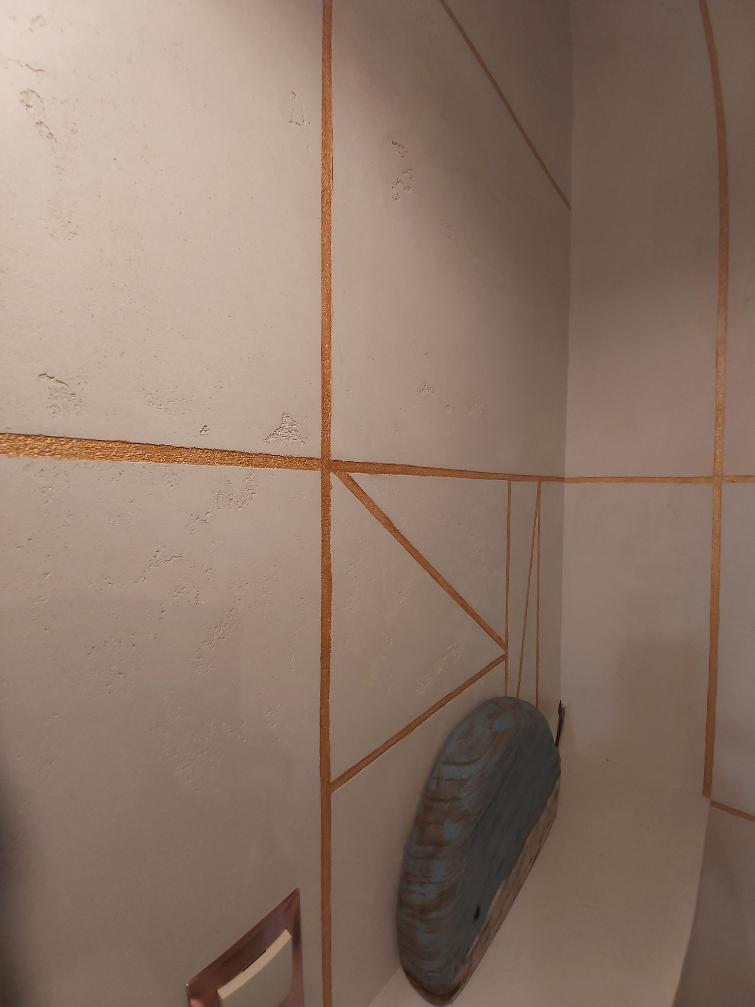 Teksturowana ściana w rogu pomieszczenia z dekoracyjnymi, złotymi liniami tworzącymi geometryczne wzory; niebieska, drewniana dekoracja przy ścianie.