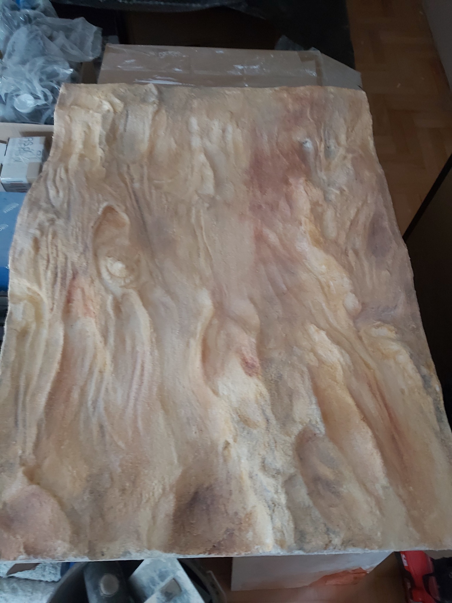 Panel dekoracyjny imitujący kamień, w odcieniach beżu i brązu, z widoczną fakturą i nierównościami, leżący na podłodze w otoczeniu narzędzi i materiałów budowlanych.