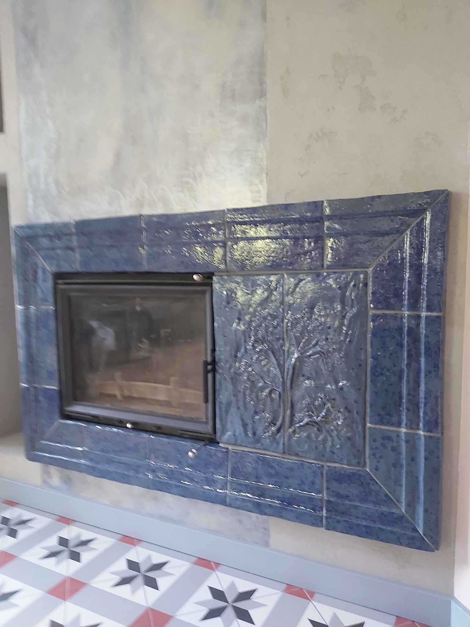 Obudowa kominka z niebieskich, ceramicznych płytek z motywem drzewa, widoczny fragment podłogi z wzorem w gwiazdy.