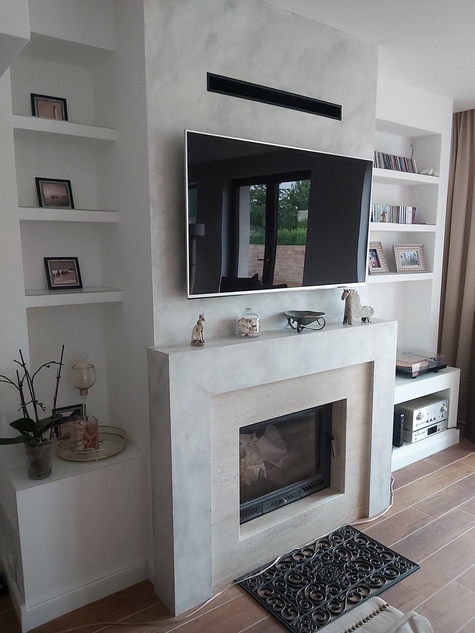 Nowoczesny salon z kominkiem obudowanym w szarym marmurze, telewizorem i białymi półkami na ścianach, uzupełniony dekoracjami.