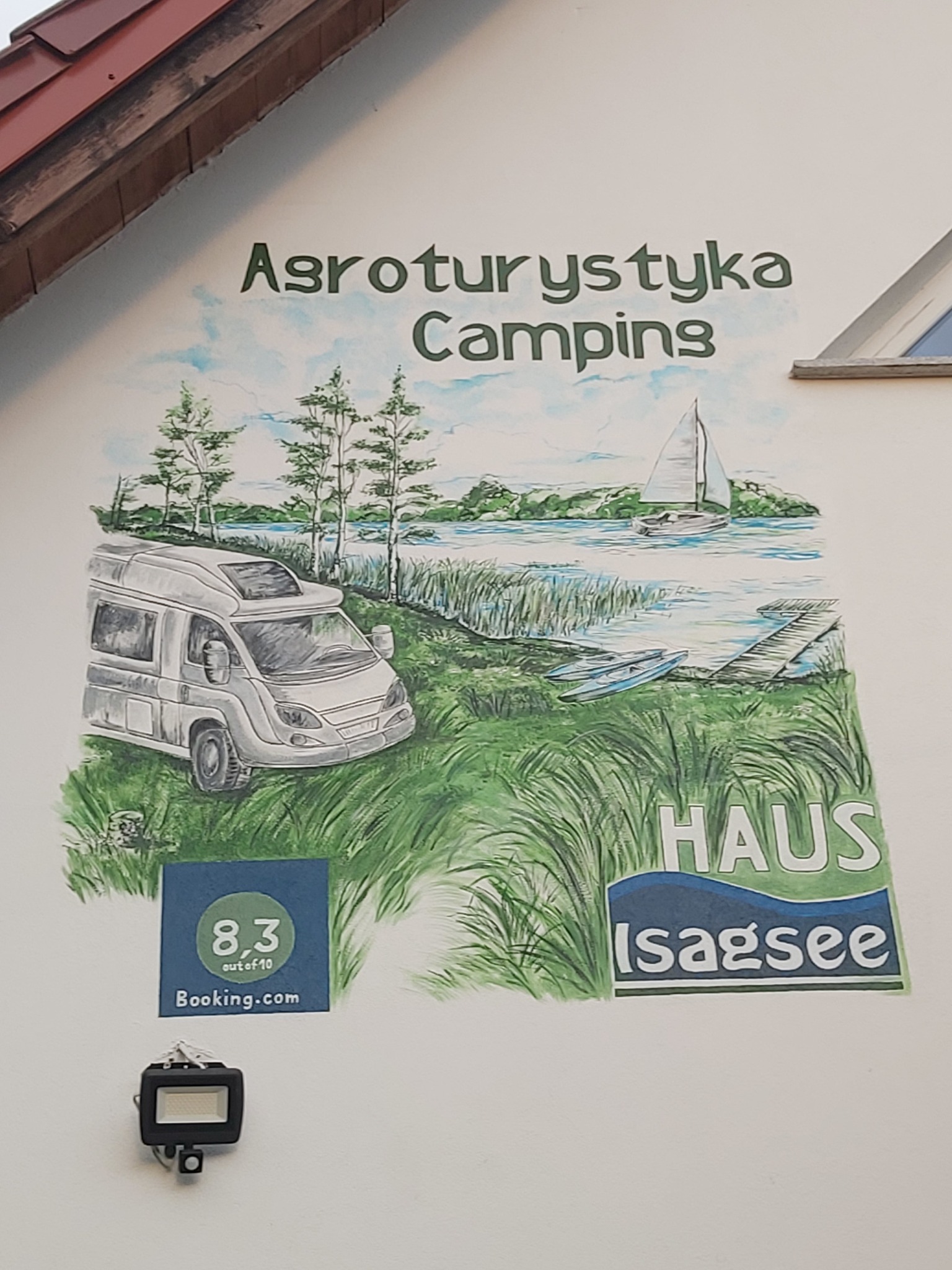 Malowidło na ścianie budynku przedstawiające krajobraz z kamperem, jeziorem, łódkami i żaglówką, z napisem 'Agroturystyka Camping' i logo 'Booking.com' z oceną 8.3/10.