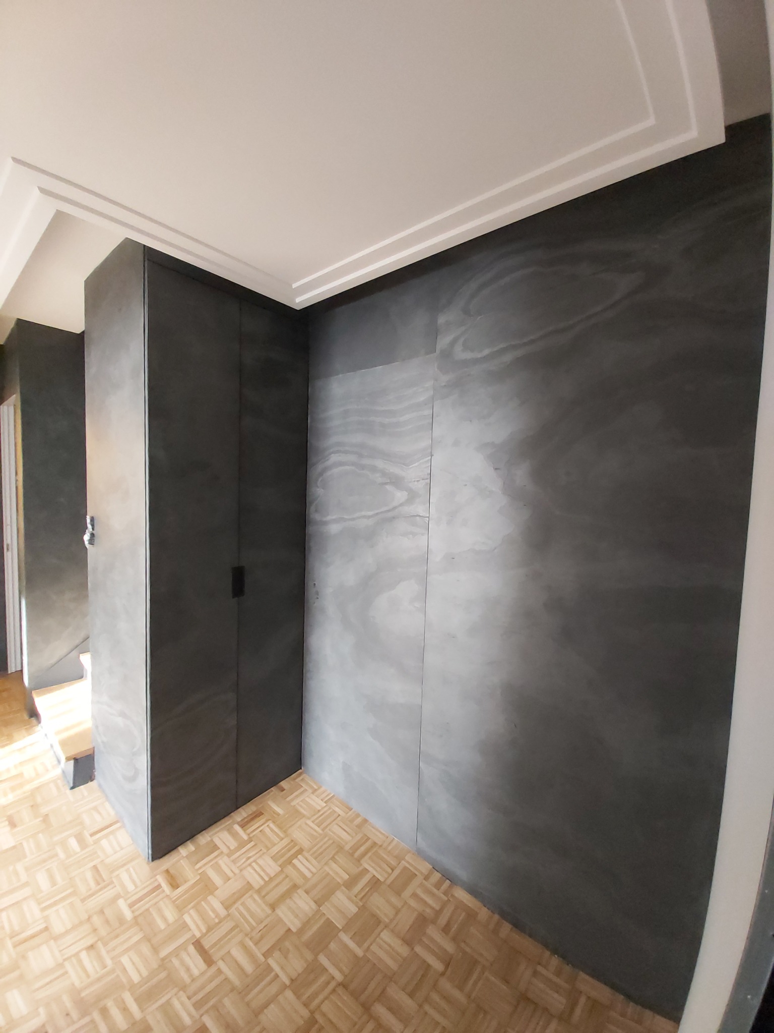 Szara ściana z efektem dekoracyjnym, zabudowana szafa w kolorze ściany, parkiet w jodełkę, białe listwy sufitowe.