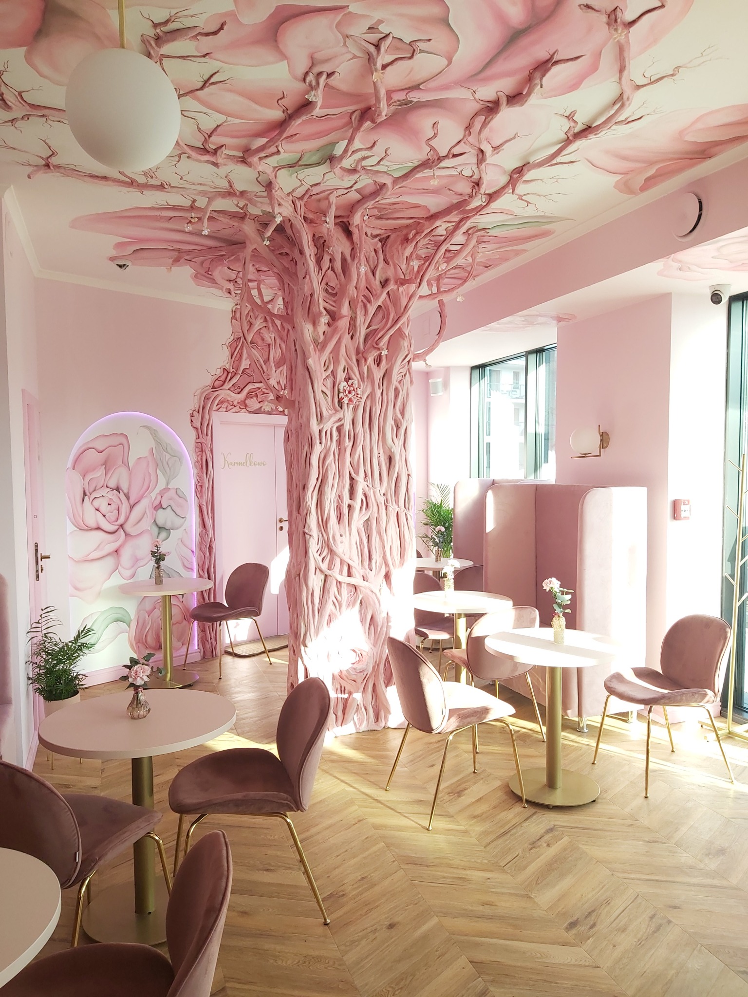 Różowe wnętrze kawiarni z dekoracyjnym drzewem wyrastającym z podłogi i rozciągającym się po suficie, okrągłe stoliki z welurowymi krzesłami na złotych nogach, parkiet ułożony w jodełkę.