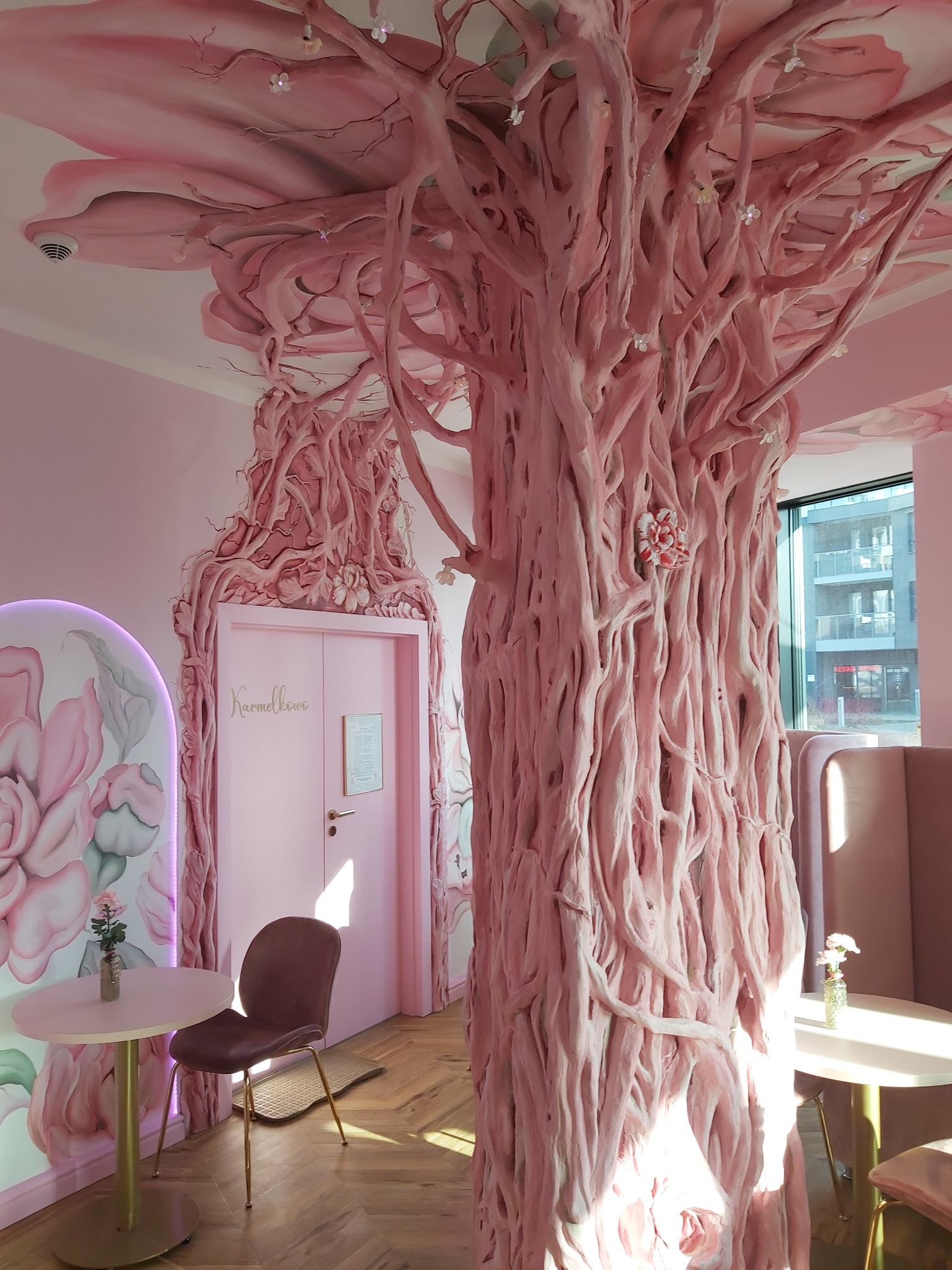 Różowe wnętrze kawiarni z dekoracyjnym drzewem oplatającym kolumnę i sufit, różowe ściany, drzwi z napisem 'Karmelkowe', stoliki z kwiatami i wygodne siedziska.