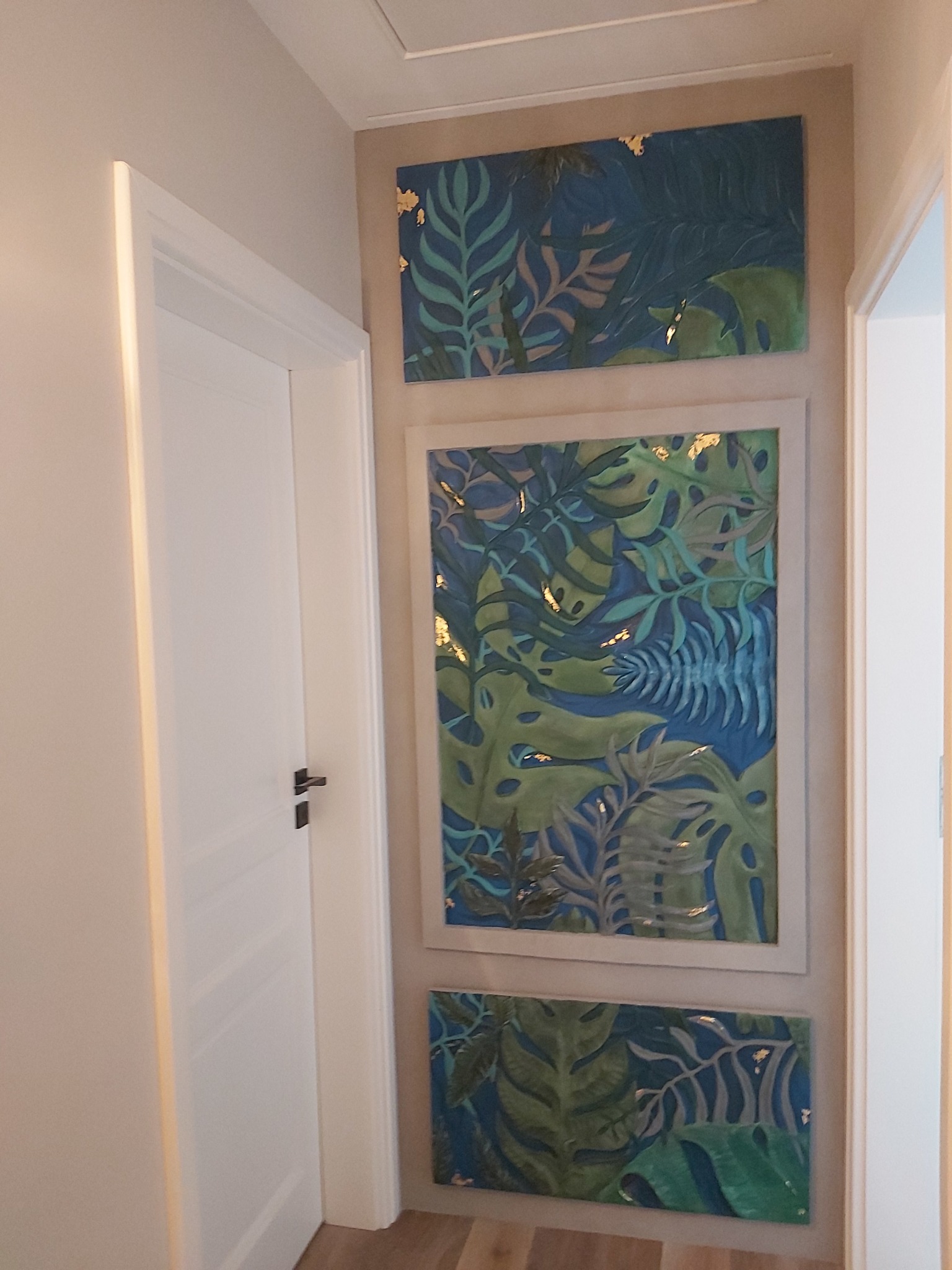 Korytarz z trzema obrazami z motywem tropikalnych liści w odcieniach zieleni i błękitu, z elementami złota, wiszącymi na beżowej ścianie obok białych drzwi.
