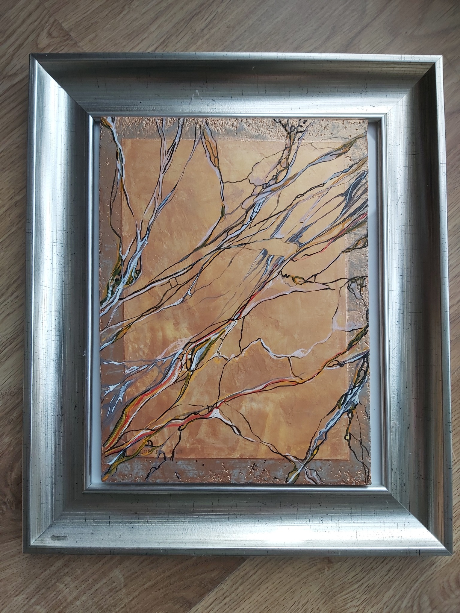 Abstrakcyjny obraz z motywem gałęzi na pomarańczowym tle, oprawiony w srebrną ramę, leżący na drewnianej podłodze.