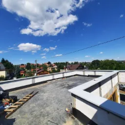 Niedokończony płaski dach z białymi obróbkami blacharskimi, widok na zabudowę jednorodzinną i błękitne niebo z chmurami.