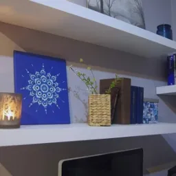 Dwie białe półki zamontowane na szarej ścianie, na których stoją książki, świeca w szklanym lampionie z motywem lasu i obraz z niebieskim tłem i białym wzorem mandali.