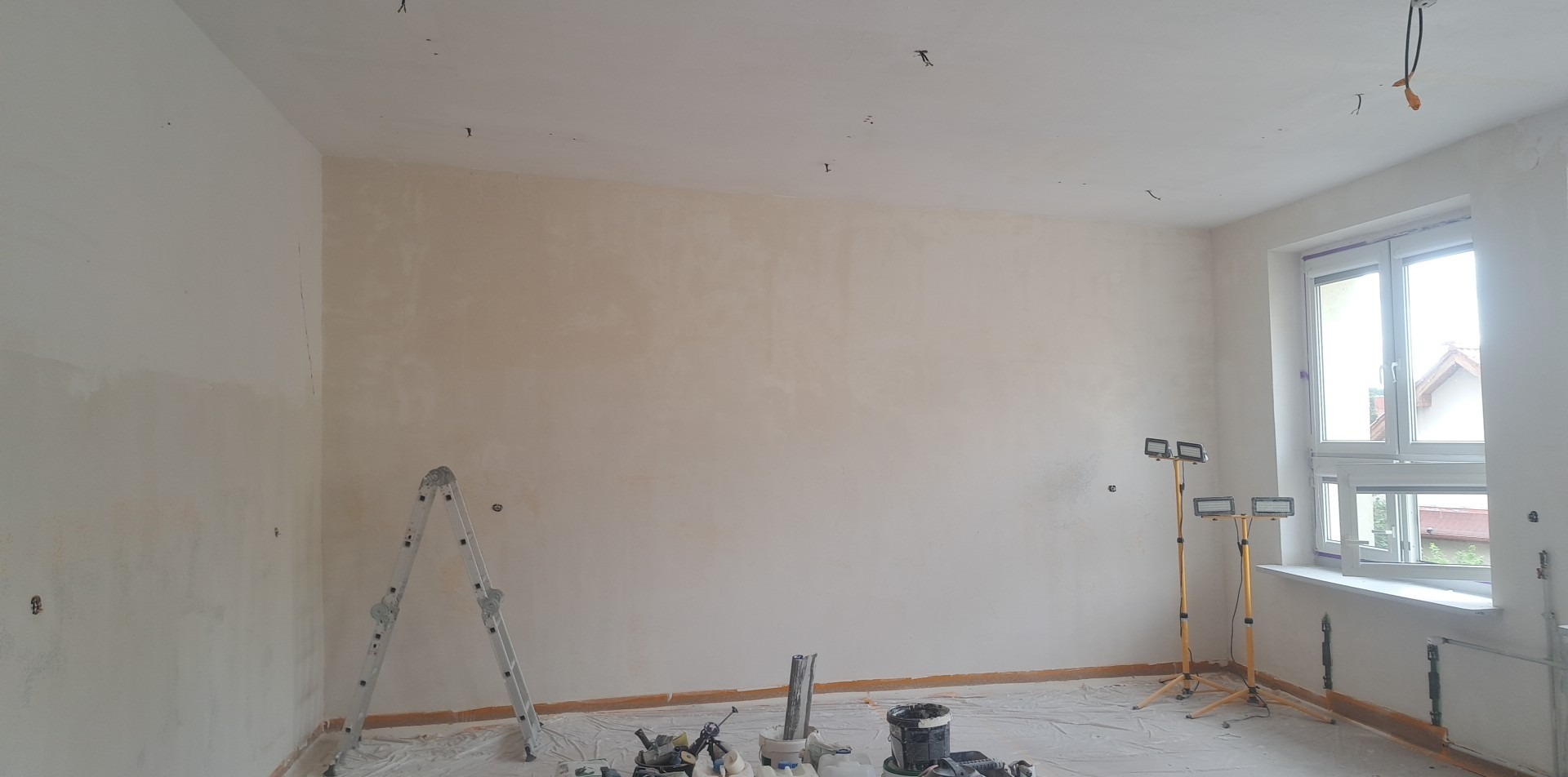 Pokój w trakcie prac wykończeniowych: ściany pokryte gładzią, widoczne narzędzia malarskie, drabina i lampy halogenowe na statywach, podłoga zabezpieczona folią malarską.