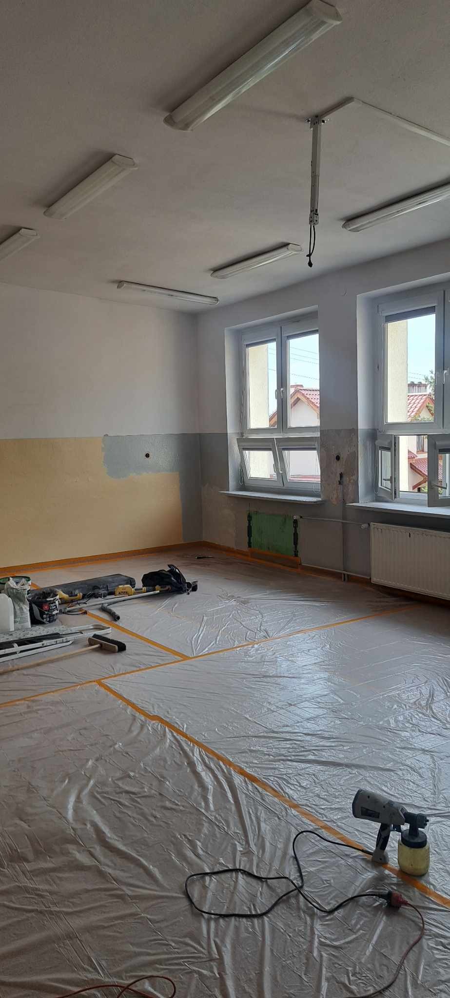 Przygotowane pomieszczenie do malowania z użyciem pistoletu natryskowego, podłoga zabezpieczona folią, widoczne narzędzia i materiały malarskie przy ścianie.
