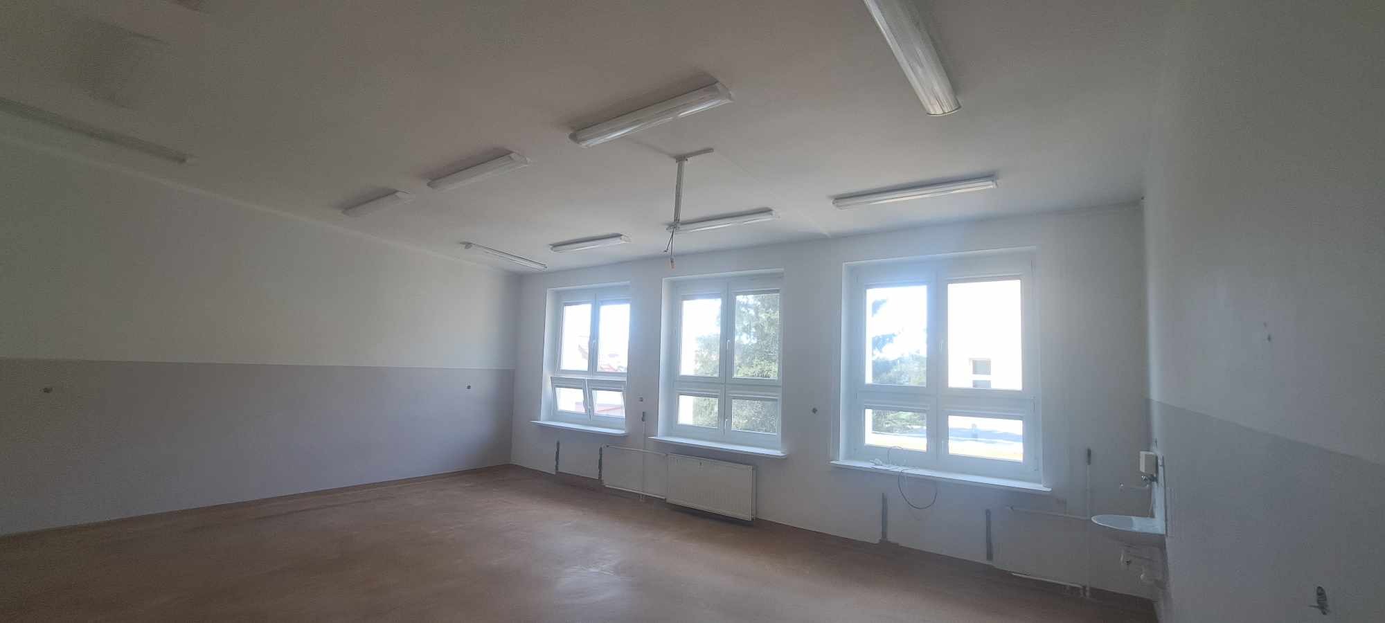 Puste pomieszczenie z trzema oknami, pomalowane na biało z szarym pasem na dole ścian, brązowa podłoga, oświetlenie sufitowe