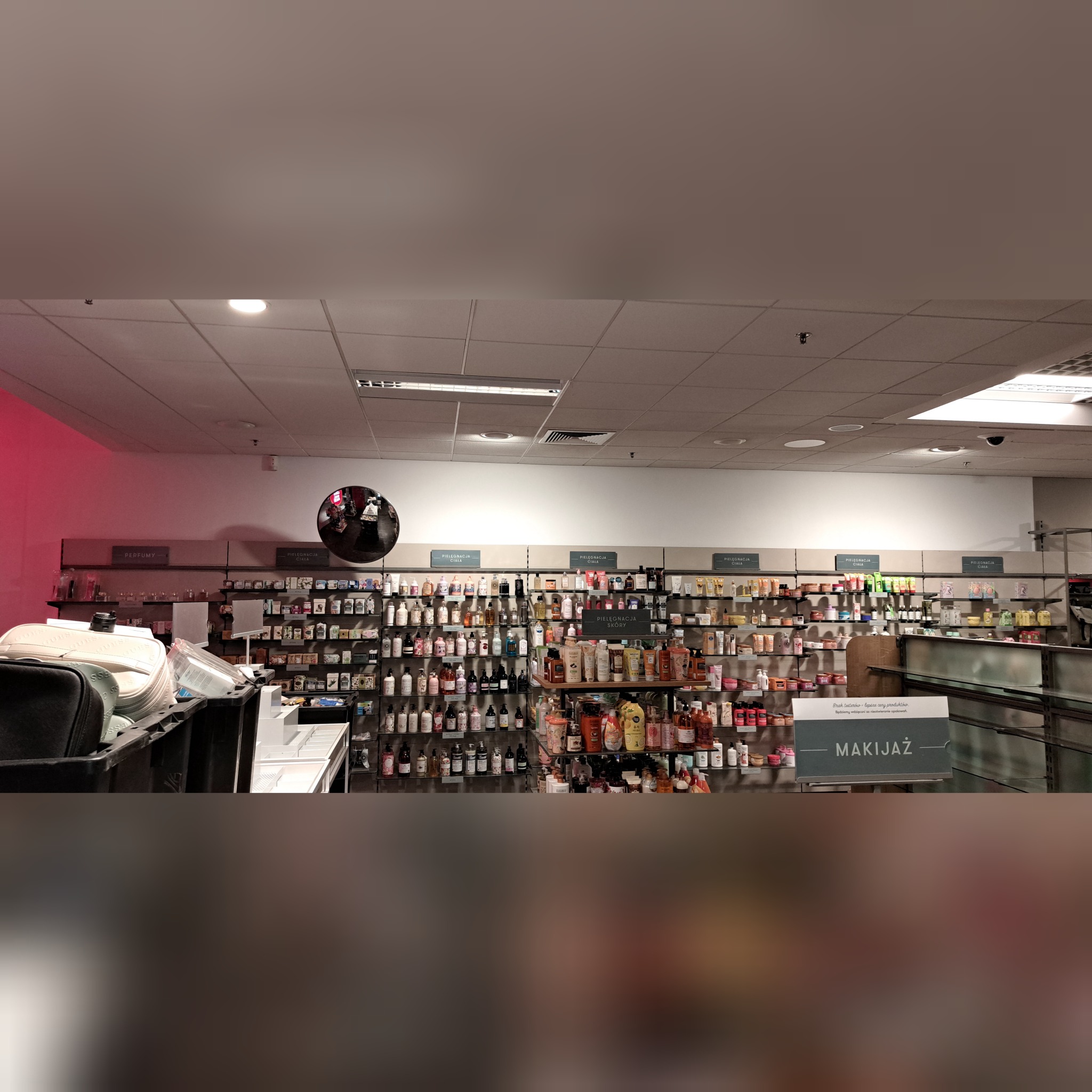 Wnętrze sklepu z kosmetykami i perfumami, półki uginają się pod ciężarem produktów do pielęgnacji ciała, skóry i makijażu, widoczny znak 'MAKIJAŻ', odbicie w lustrze sferycznym.