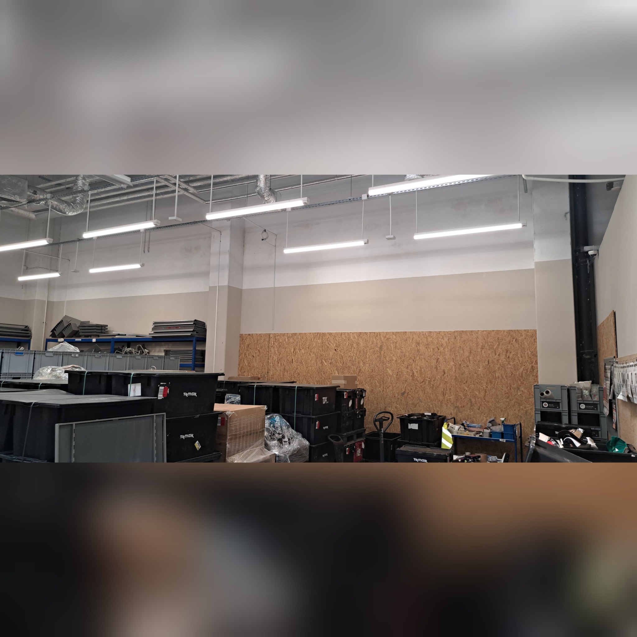 Wnętrze magazynu lub hali z widocznymi regałami, skrzyniami transportowymi i płytą OSB na ścianie, oświetlone rzędami lamp jarzeniowych.
