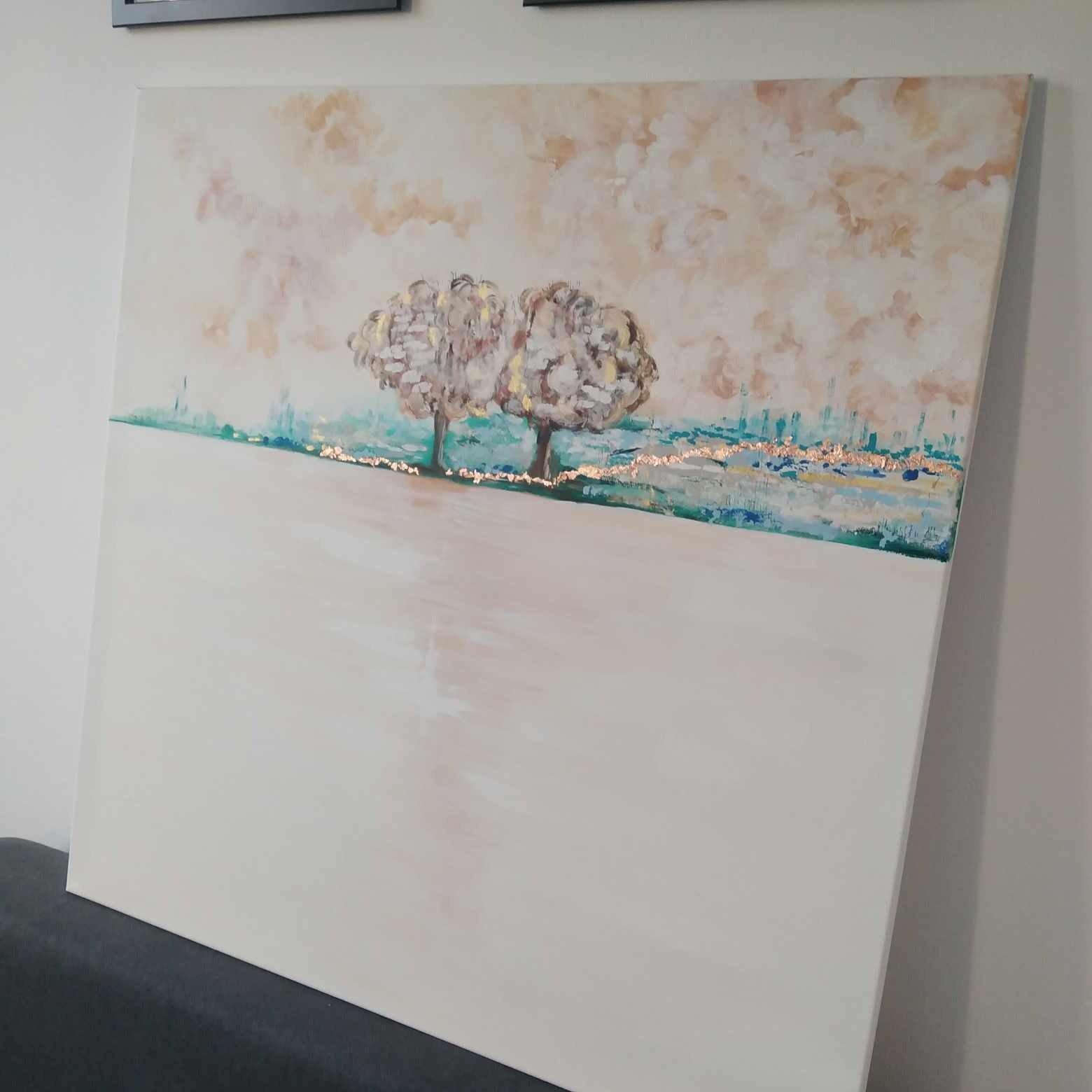 Abstrakcyjny obraz przedstawiający dwa drzewa z elementami złota na płótnie, w odcieniach beżu, bieli, zieleni i błękitu, umieszczony na szarej sofie.