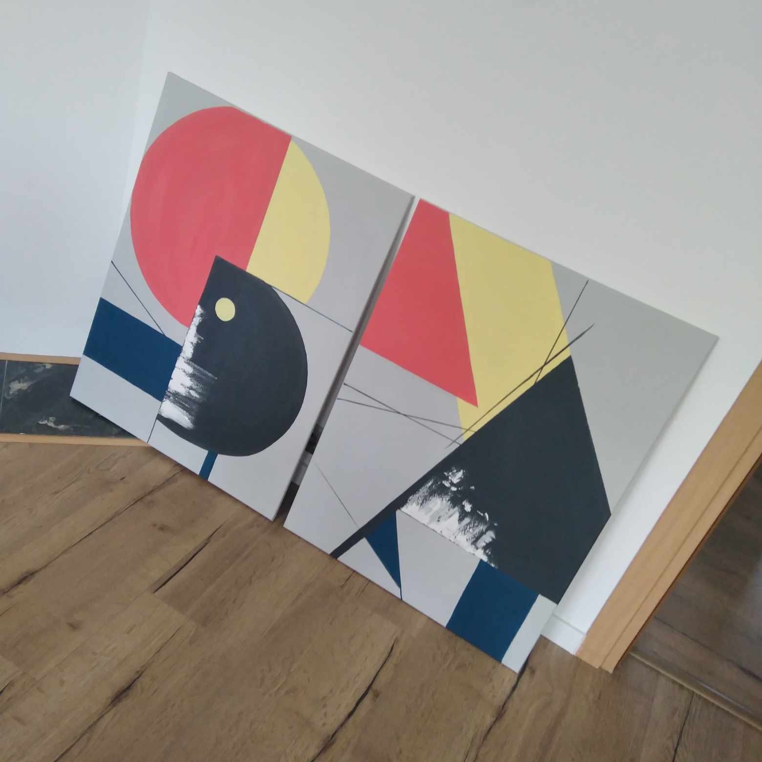 Dwa abstrakcyjne obrazy geometryczne w odcieniach czerwieni, żółci, szarości i granatu, oparte o ścianę na drewnianej podłodze, z widocznymi cienkimi liniami grafitu.