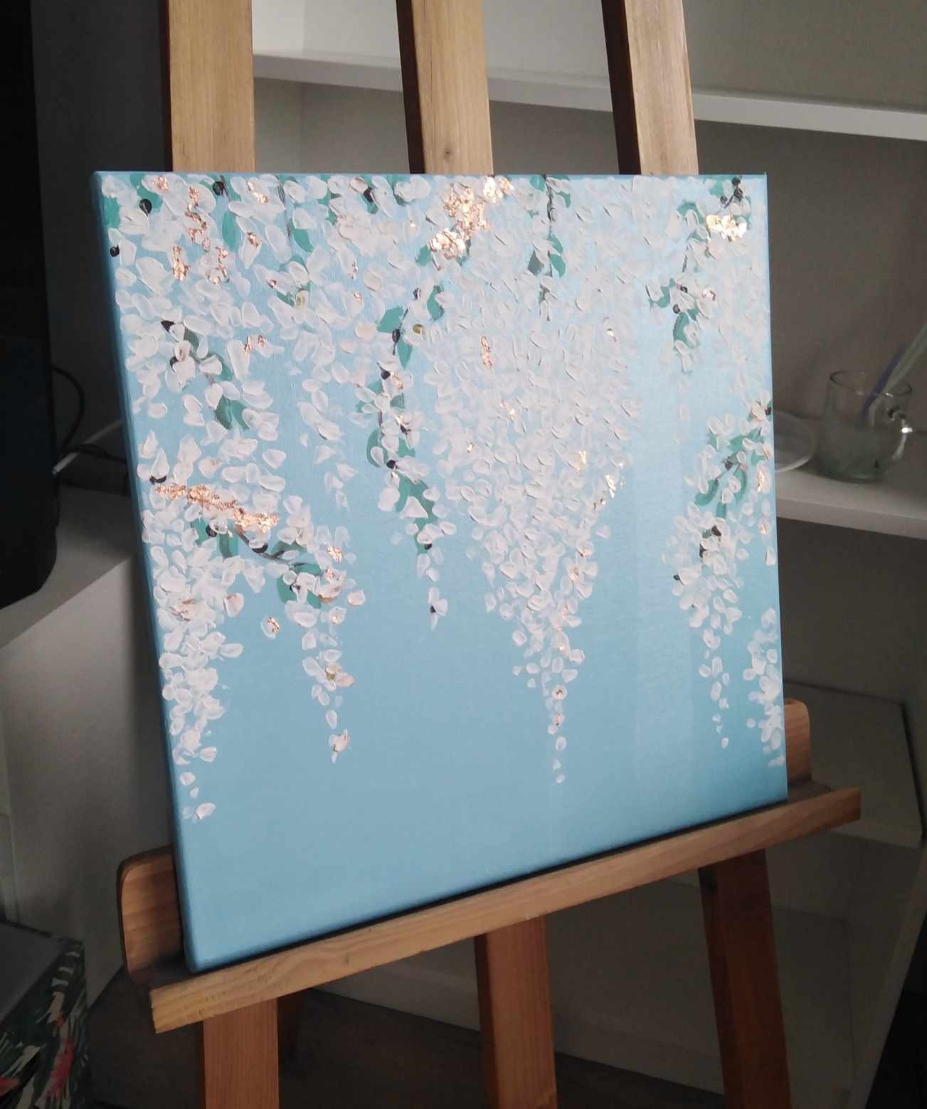 Obraz przedstawiający białe kwiaty z akcentami złota na błękitnym tle, umieszczony na drewnianej sztaludze, widoczny z boku.