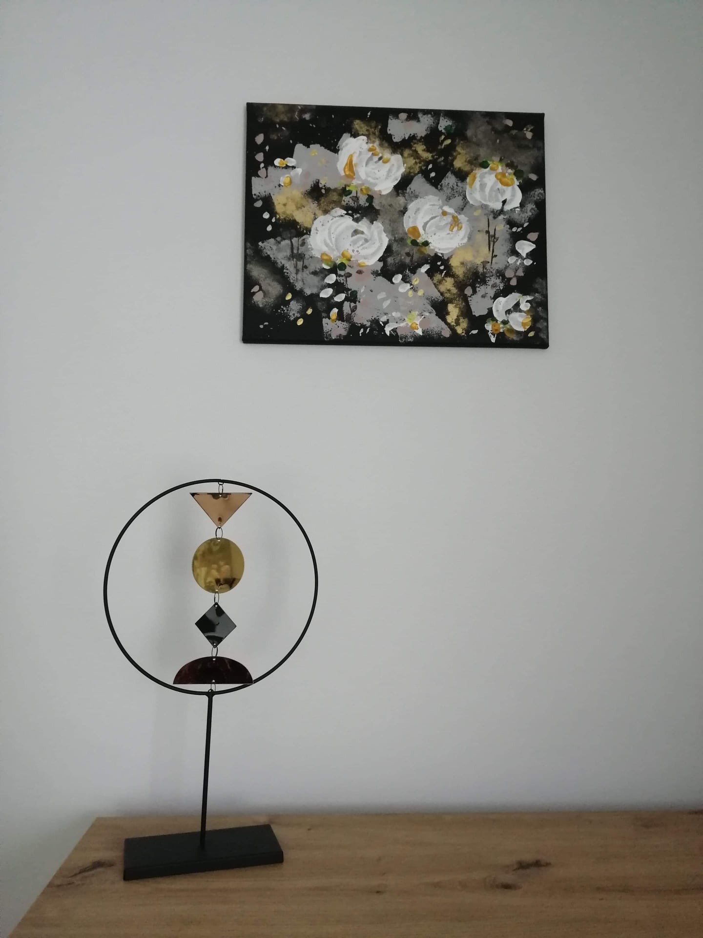 Abstrakcyjny obraz z białymi kwiatami nad minimalistyczną rzeźbą z metalu i drewna na drewnianym blacie, na tle białej ściany.