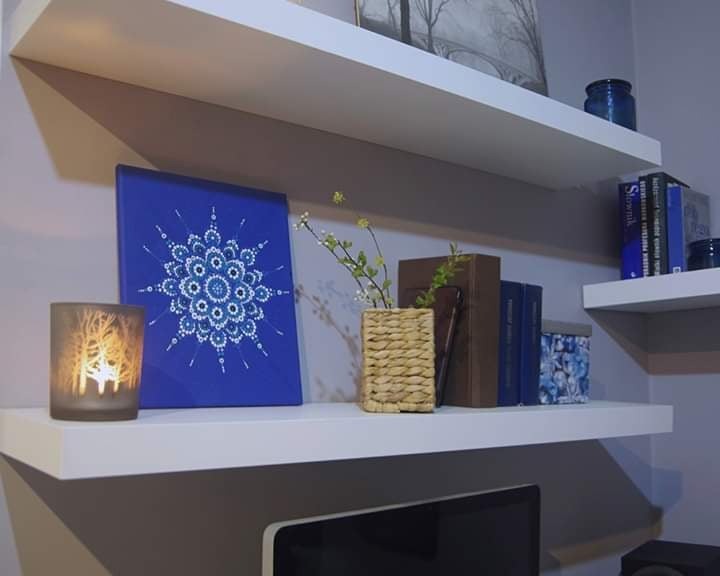 Dwie białe półki zamontowane na szarej ścianie, na których stoją książki, świeca w szklanym lampionie z motywem lasu i obraz z niebieskim tłem i białym wzorem mandali.