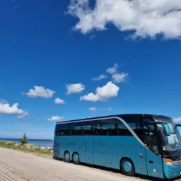 Turkusowy autokar Setra zaparkowany na poboczu z widokiem na morze i błękitne niebo z chmurami.