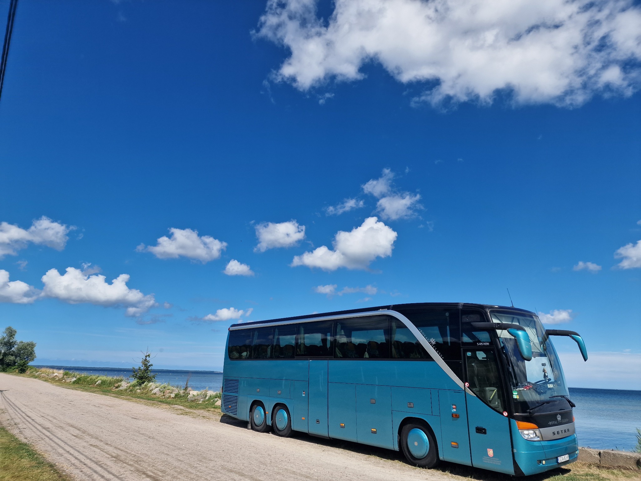 Turkusowy autokar Setra zaparkowany na poboczu z widokiem na morze i błękitne niebo z chmurami.