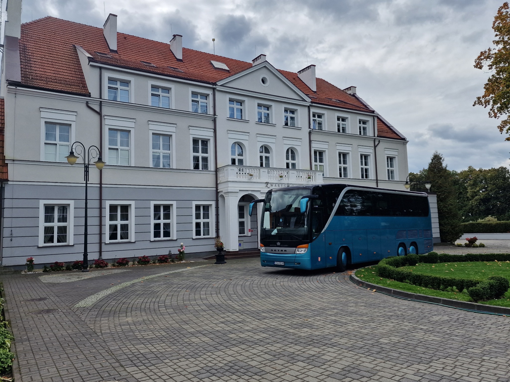 Niebieski autokar SETRA Royal Class zaparkowany przed klasycystycznym budynkiem z czerwonym dachem i balkonem, otoczony brukowanym podjazdem i zielenią.