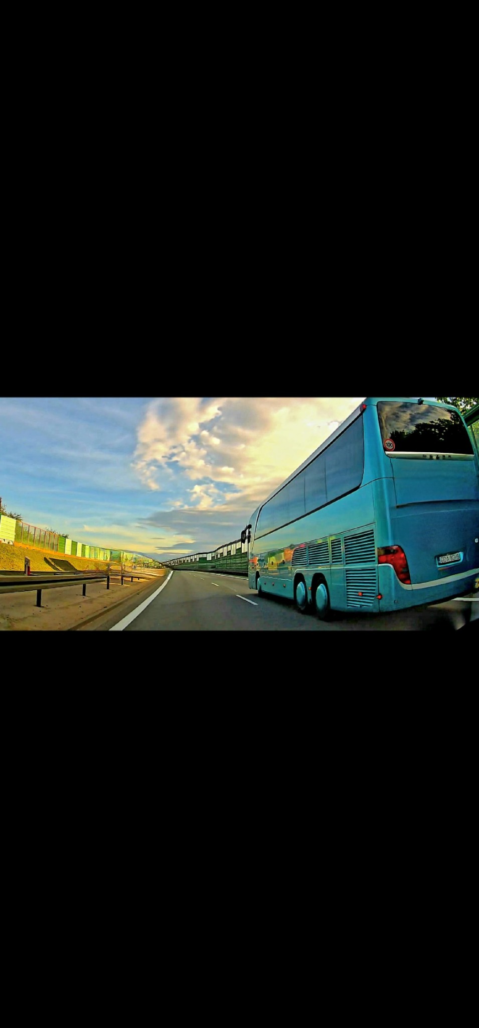 Niebieski autokar na autostradzie, widok z boku, w tle bariery dźwiękochłonne i błękitne niebo z chmurami.