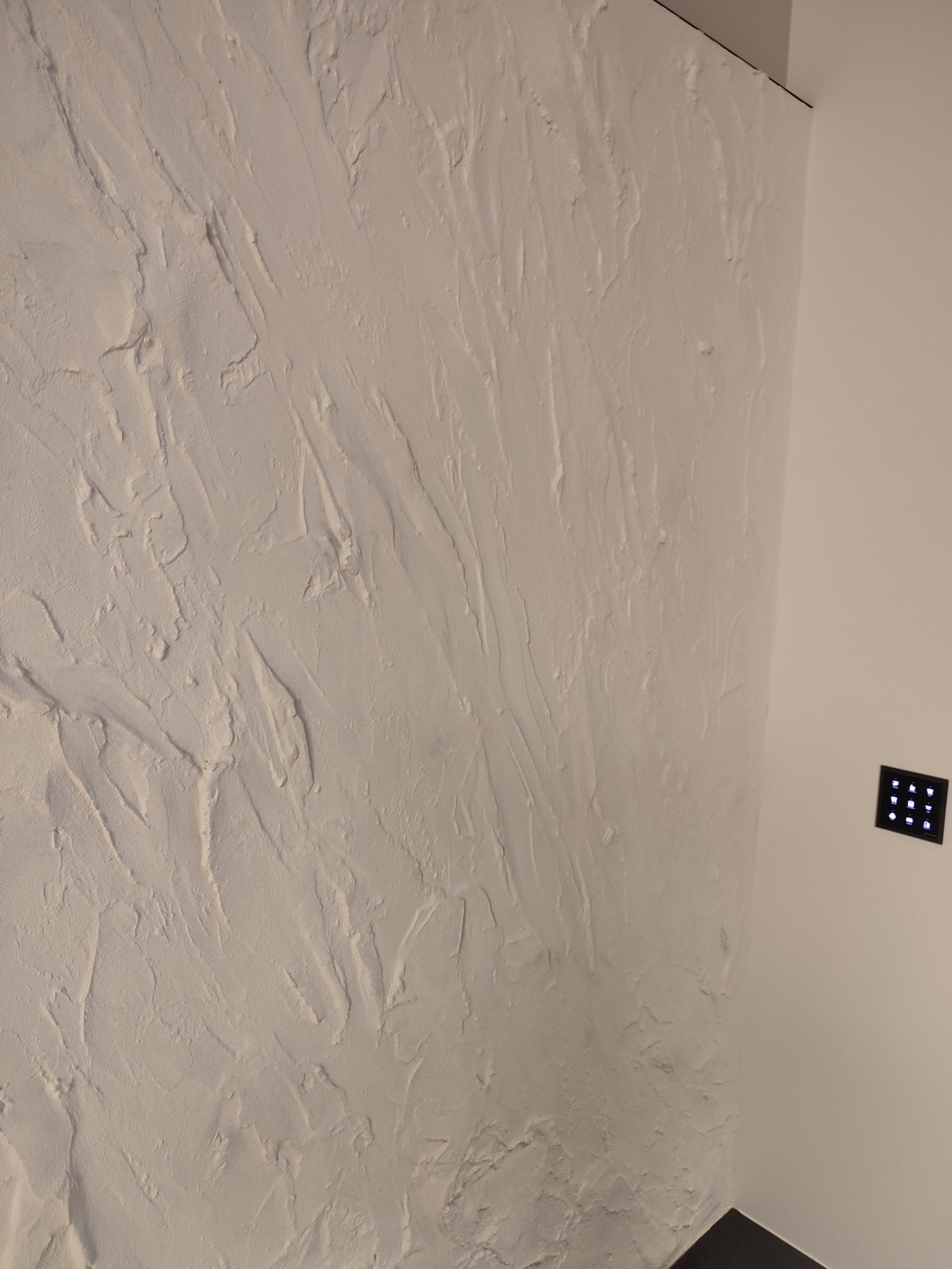 Biała ściana z wyraźną, nieregularną fakturą szpachlowania, kontrastująca z gładką ścianą i czarnym panelem sterowania oświetleniem.