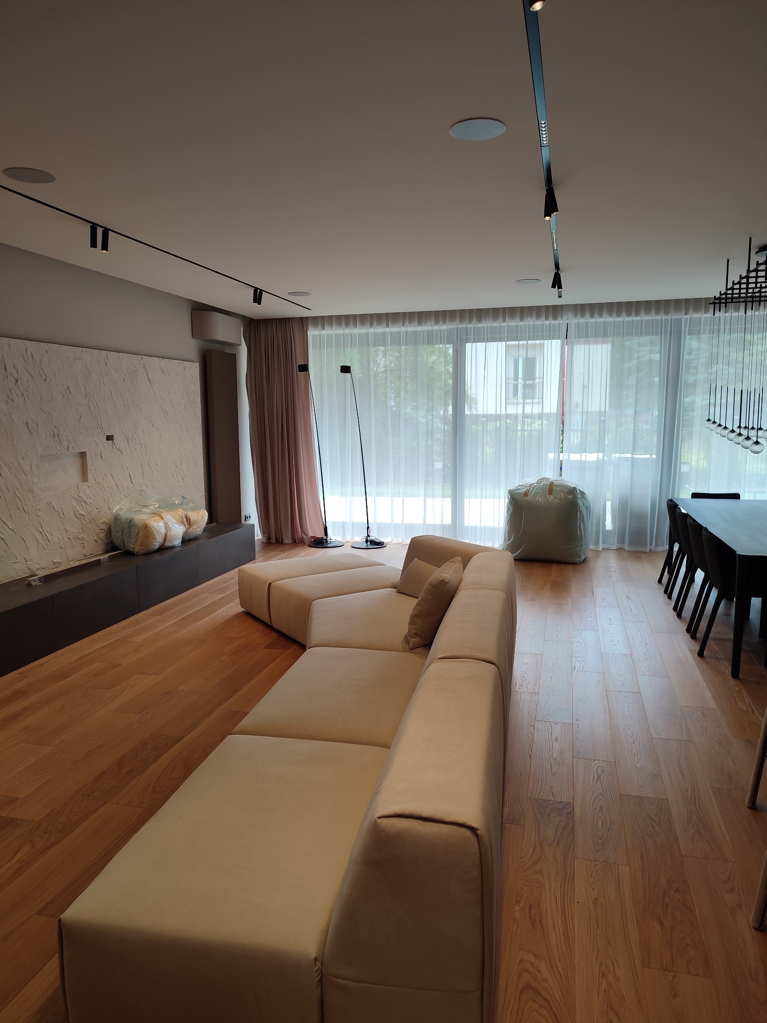 Jasne, przestronne wnętrze salonu z drewnianą podłogą, beżową sofą modułową, ścianą z dekoracyjną fakturą i dużymi oknami zasłoniętymi delikatnymi firanami. Widoczne nowoczesne oświetlenie sufitowe...