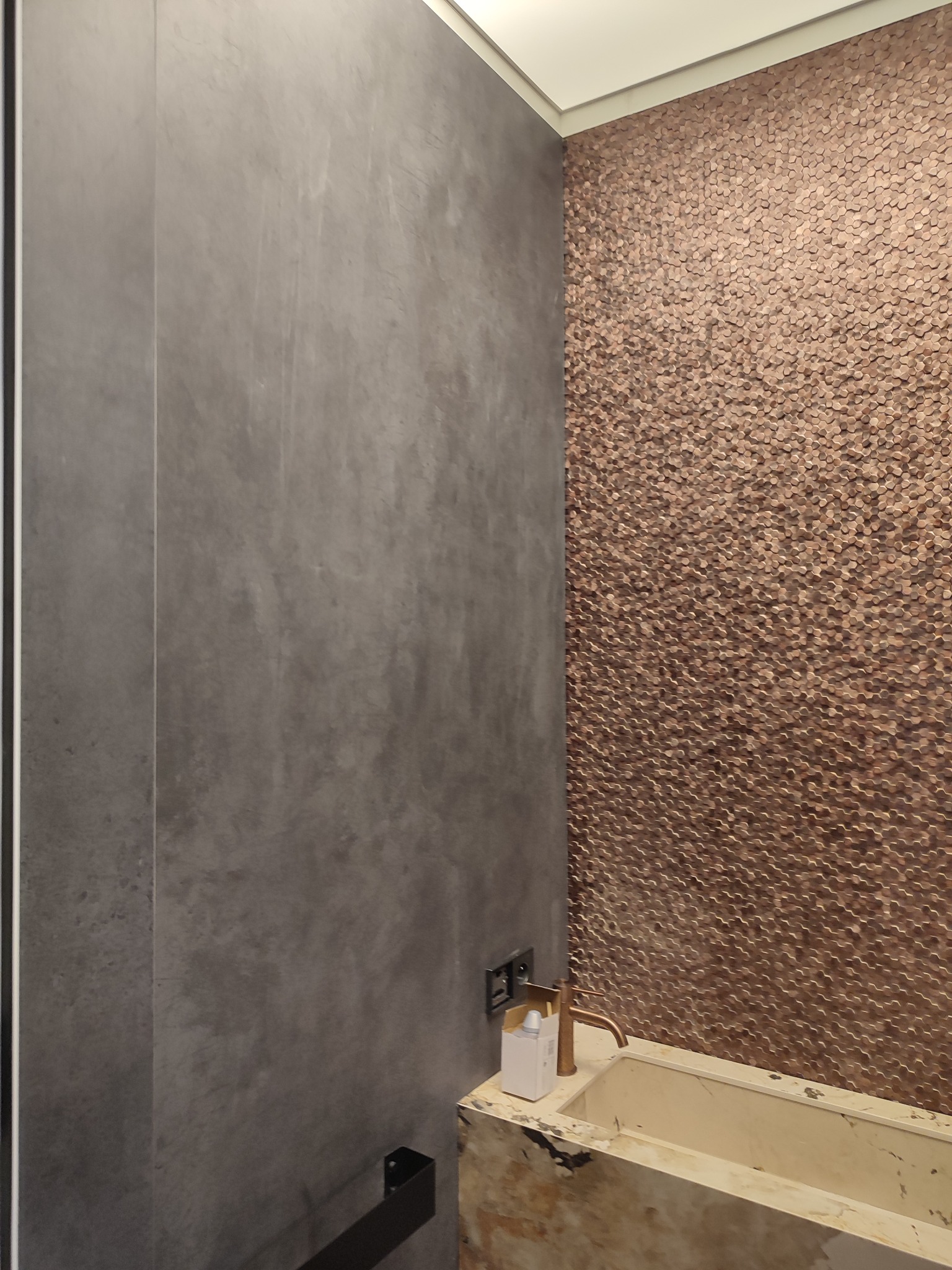 Dekoracyjna ściana w łazience w Warszawie, wykończona mozaiką w odcieniach miedzi, kontrastująca z szarą, gładką powierzchnią ściany obok umywalki z marmurowym blatem i miedzianą armaturą.