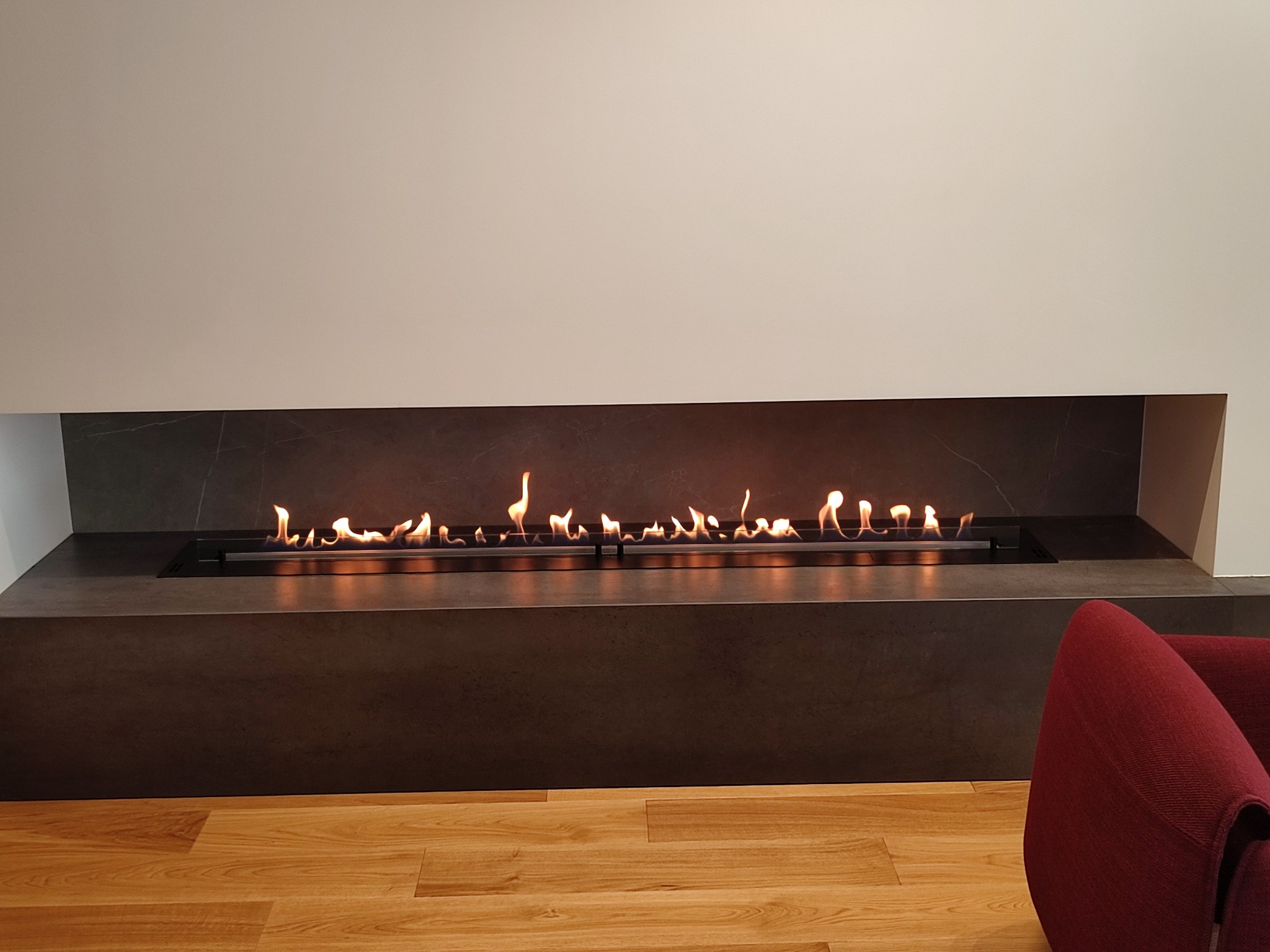 Wbudowany biokominek z czarną obudową i płomieniami, widoczny fragment drewnianej podłogi i czerwonego fotela.