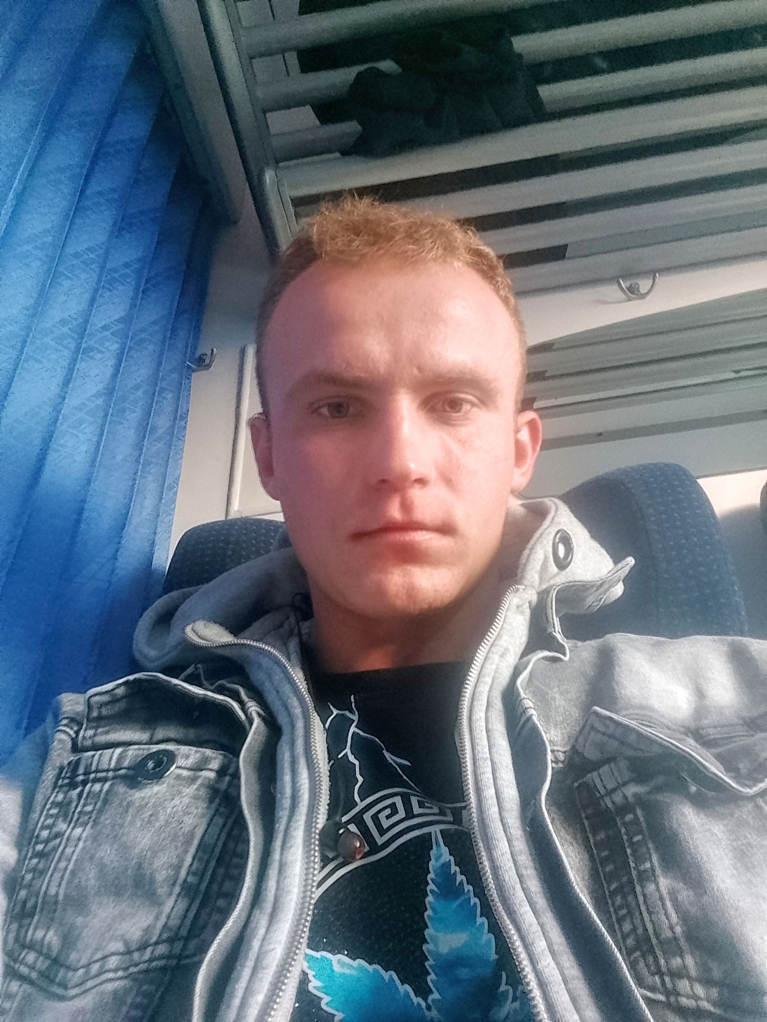 Autoportret mężczyzny w szarej kurtce dżinsowej z kapturem i koszulce z nadrukiem, siedzącego na siedzeniu w pociągu z niebieskimi zasłonami w tle.