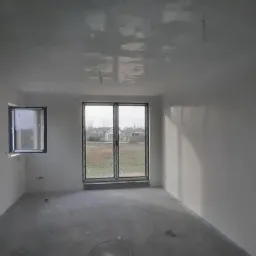 Pokój w trakcie remontu, ściany i sufit po nałożeniu pierwszej warstwy białej gładzi, widoczne okno i drzwi balkonowe z widokiem na zewnątrz, szary betonowy podkład na podłodze.
