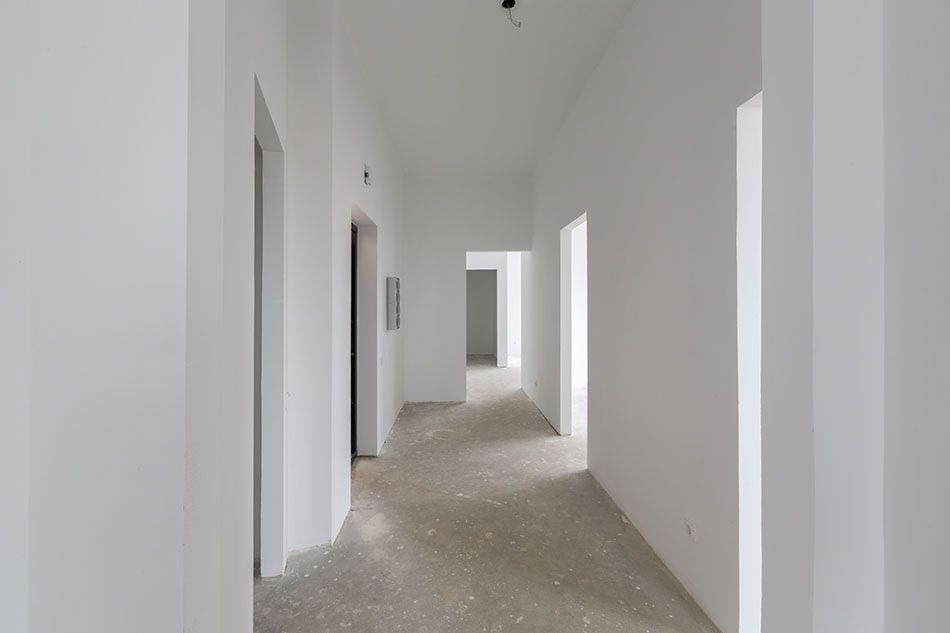 Długi korytarz w stanie surowym z białymi ścianami po szpachlowaniu i przygotowaniu do malowania, widoczne otwory drzwiowe i surowa podłoga.