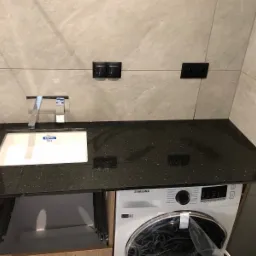 Czarny brokatowy blat łazienkowy z umywalką wpuszczaną, chromowaną baterią i pralką pod spodem; szare płytki na ścianie i czarne gniazdka elektryczne.