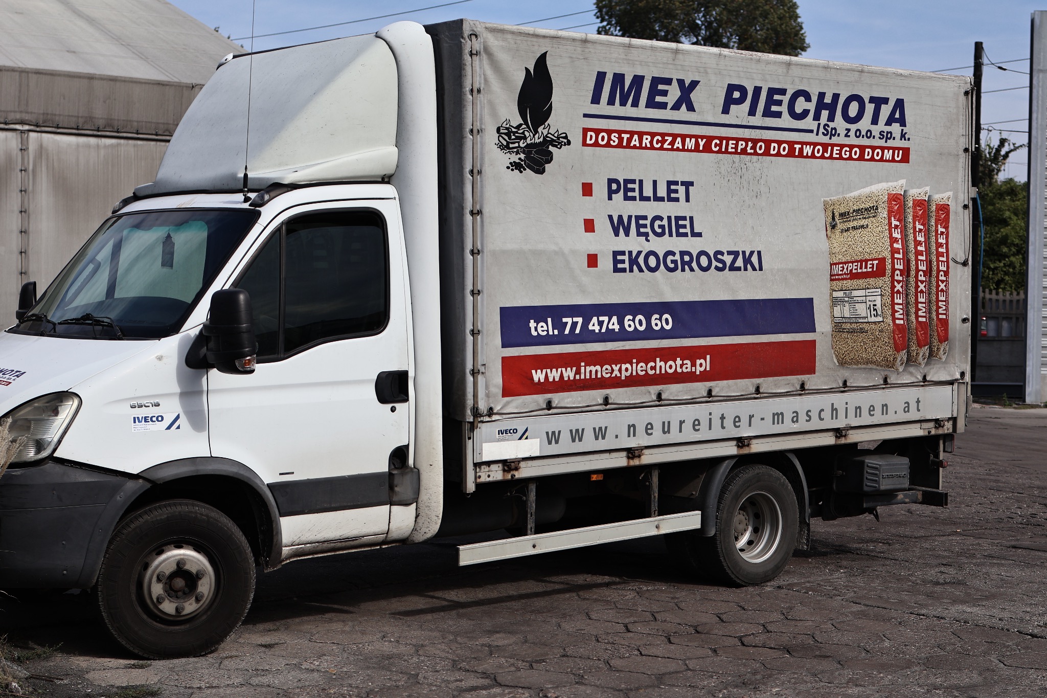 Biała furgonetka dostawcza z reklamą firmy IMEX PIECHOTA oferującej pellet, węgiel i ekogroszek, zaparkowana na brukowanej powierzchni.