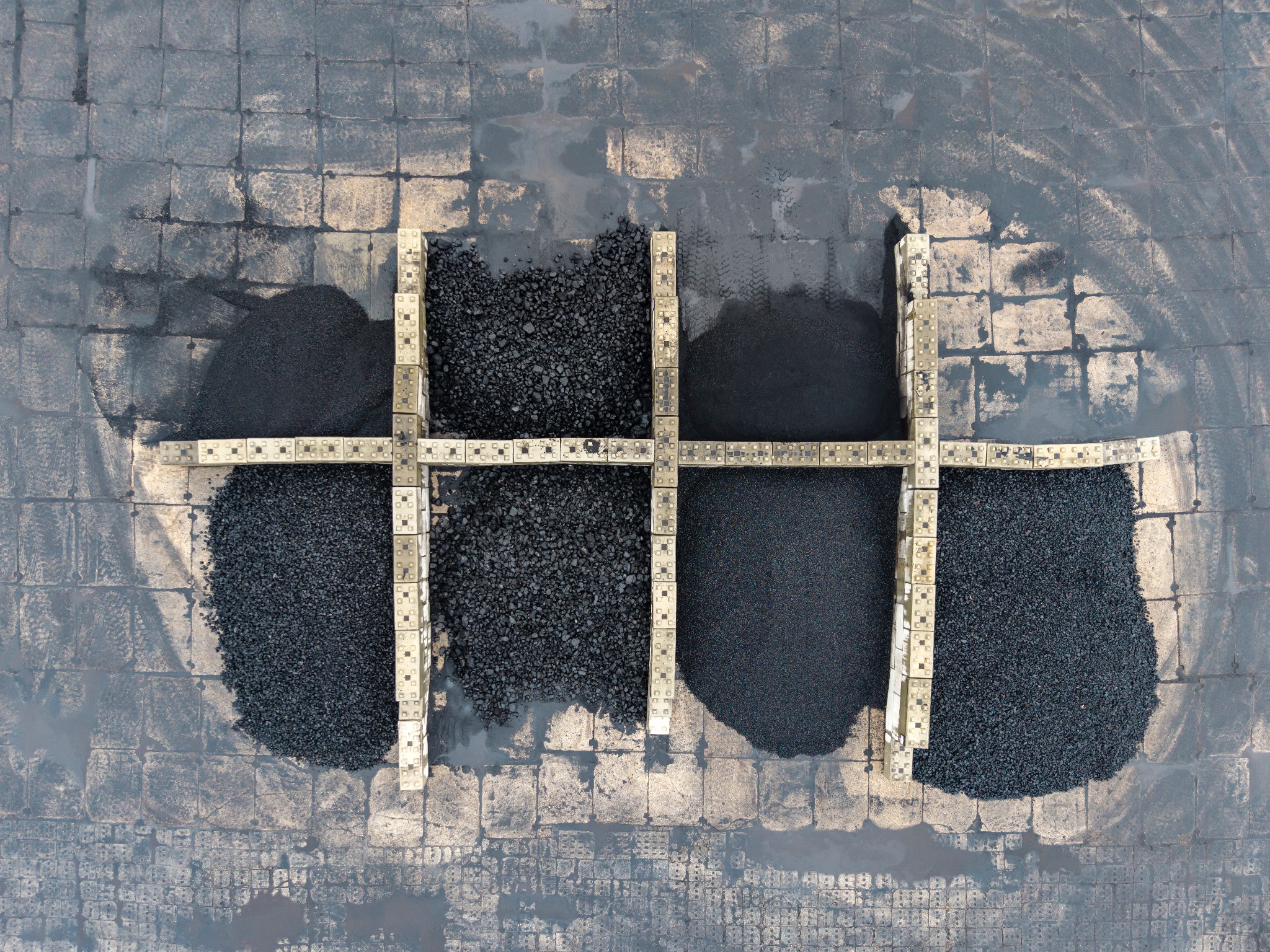 Widok z góry na cztery pryzmy czarnego opału o różnej grubości ziarna, oddzielone betonowymi krawężnikami na placu wyłożonym kostką brukową.