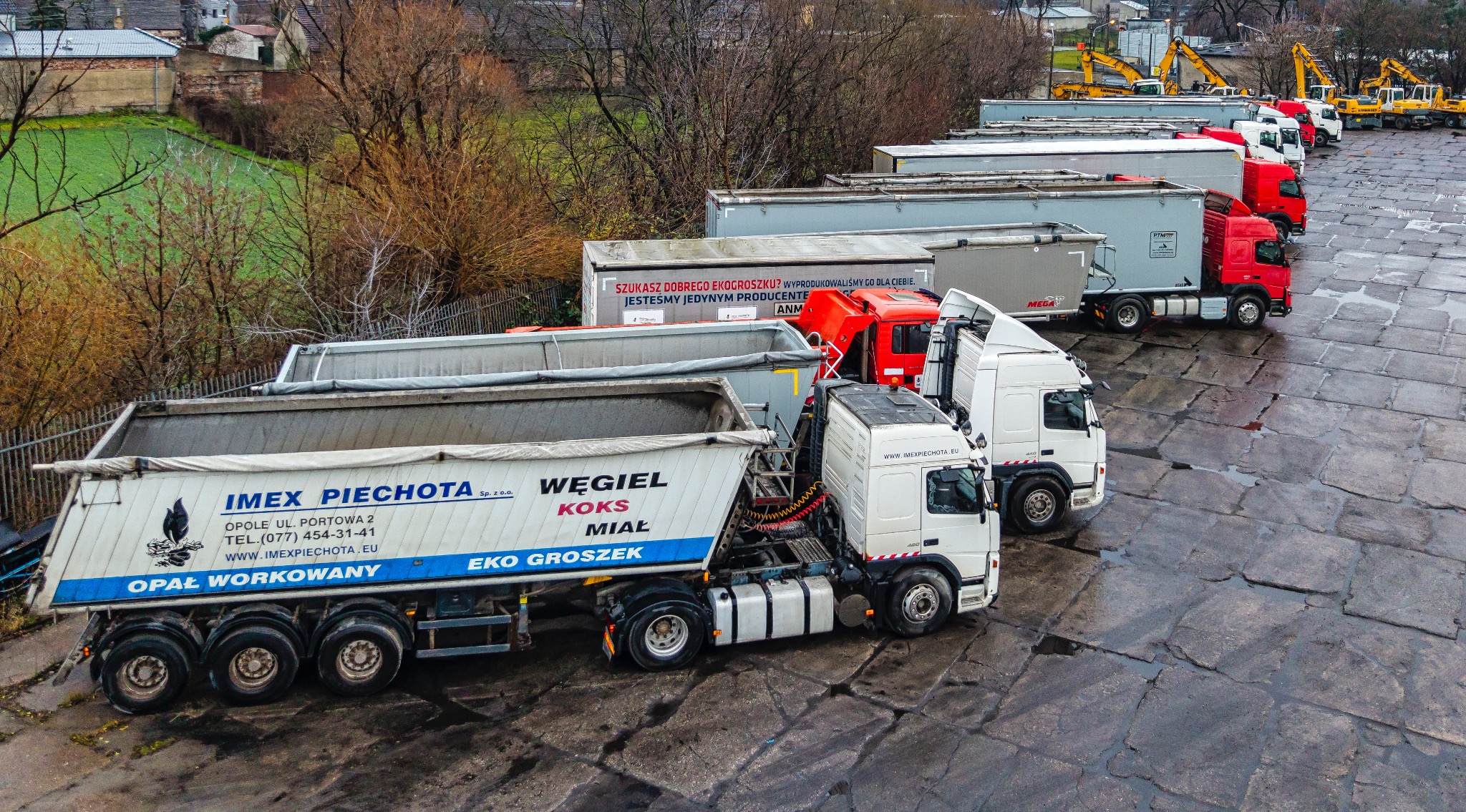 Flota ciężarówek z naczepami wywrotkami firmy Imex Piechota z Opola, z reklamami węgla, koksu, miału i ekogroszku, zaparkowana na placu z betonowych płyt, w tle drzewa i żółte koparki.