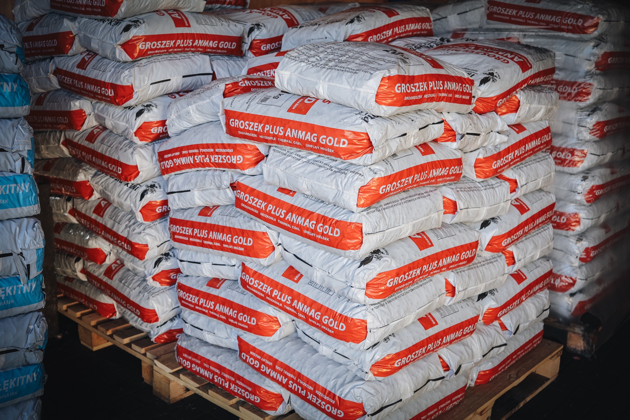 Stos palet z workami białego opału z czerwonym nadrukiem 'Groszek Plus Anmag Gold', widoczne fragmenty napisów w języku polskim i czeskim, oświetlenie magazynowe.